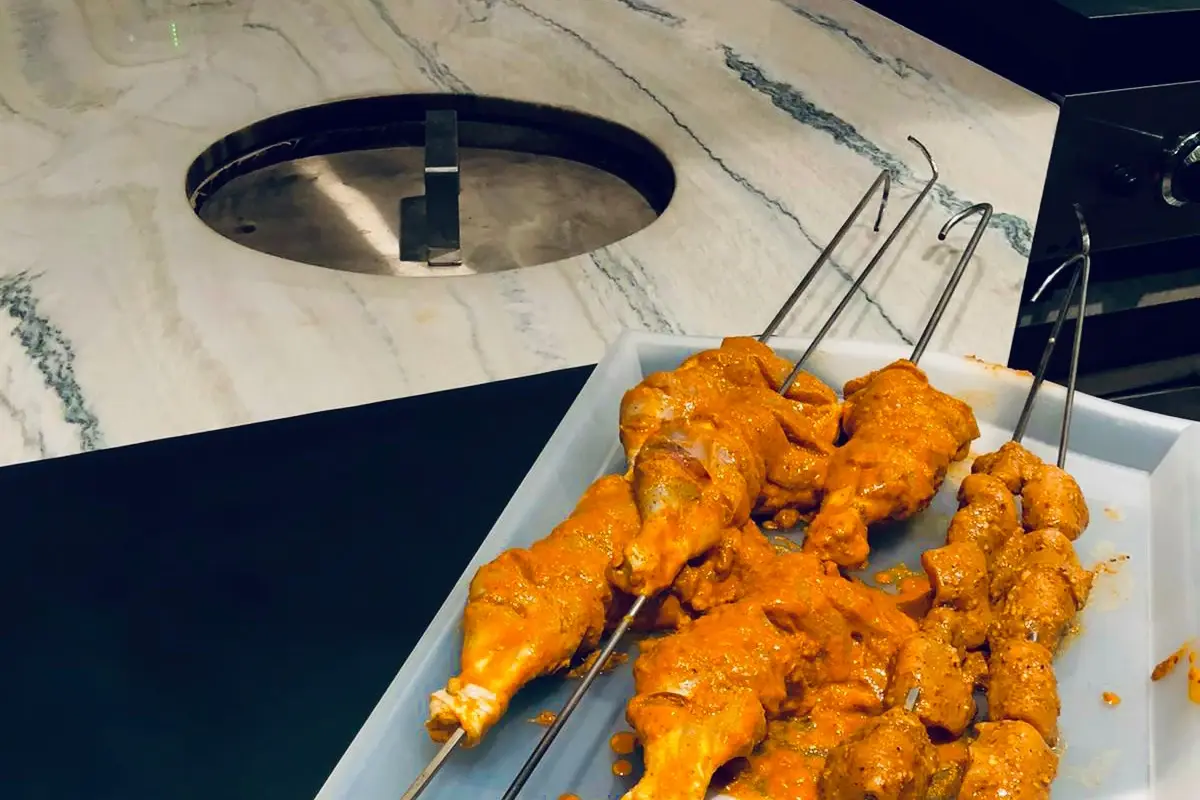 GULATI tandoor built into a marble outdoor kitchen countertop, with tandoori chicken skewers ready to cook